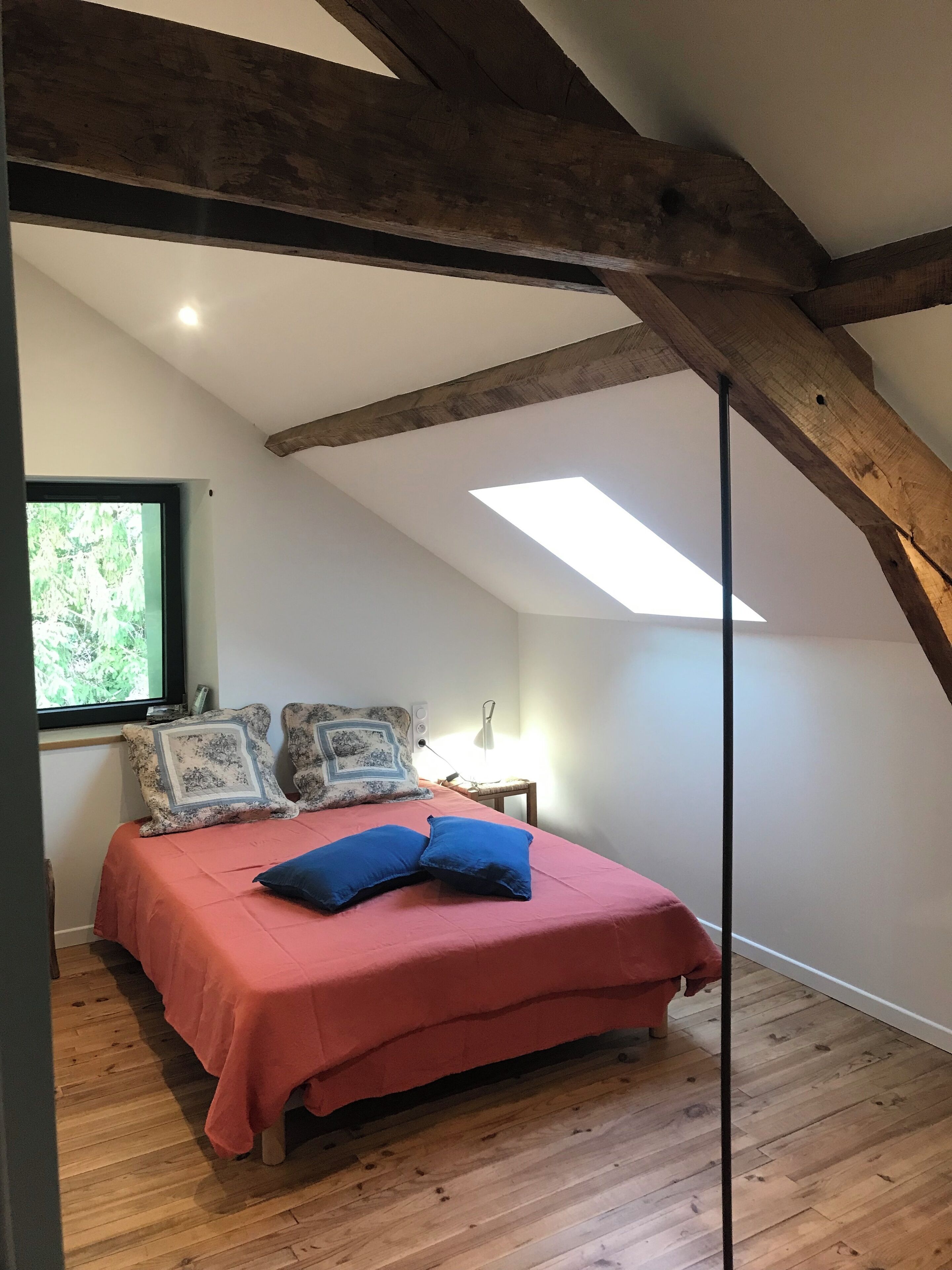 A bedroom with a sloped ceiling, wooden beams, a skylight, a bed with a red bedspread and two blue pillows, a nightstand with a lamp, and a window with a view of greenery.