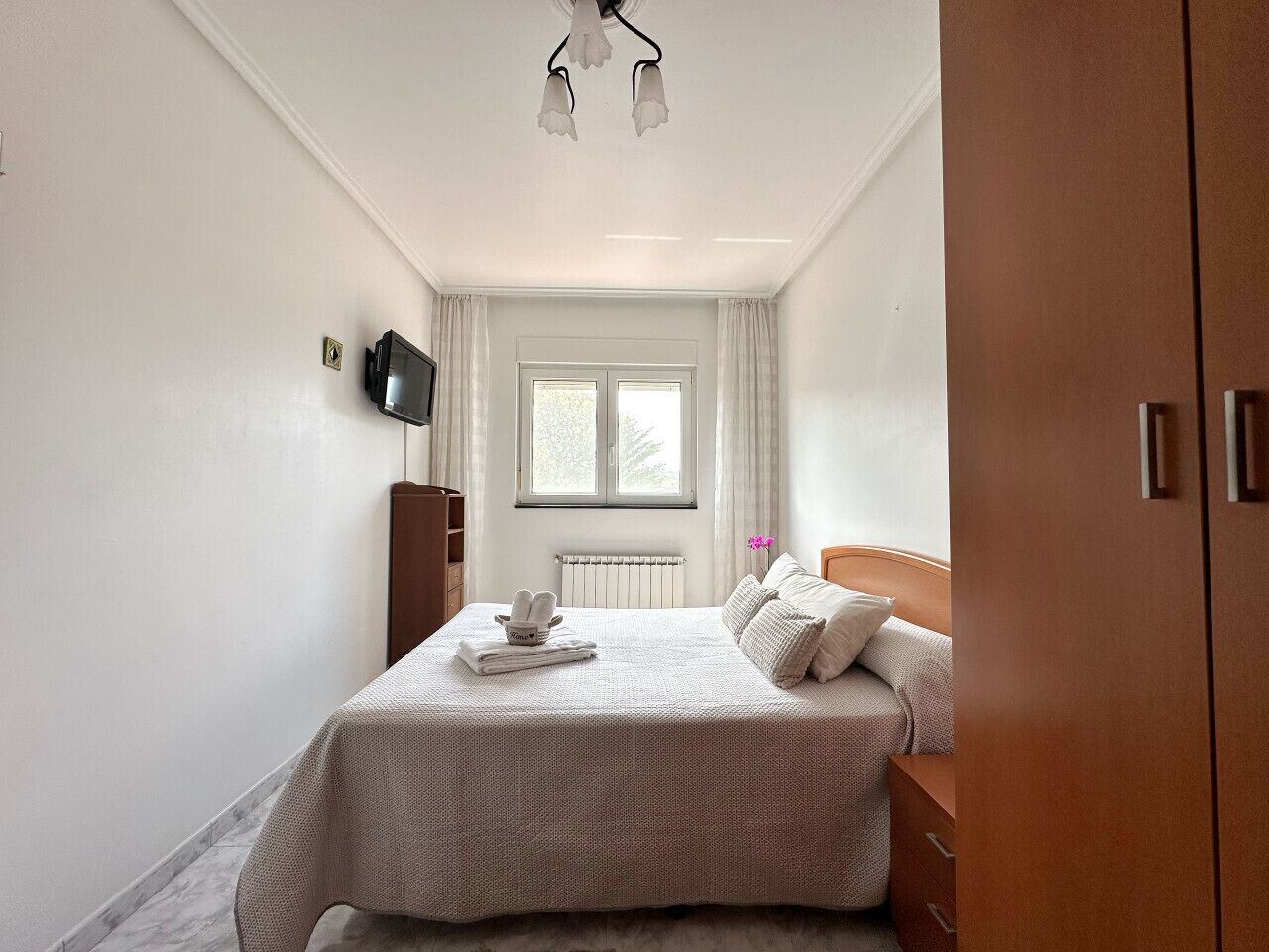 Une chambre à coucher bien rangée, avec un lit, un téléviseur fixé au mur, une fenêtre avec des rideaux et un radiateur.