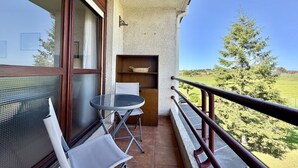 A balcony with a table and chairs, a small shelf, and a view of a green landscape.