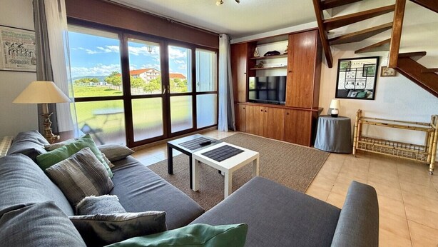 A living room with a sofa, coffee table, and a TV. There is a staircase and a view of the outdoors.