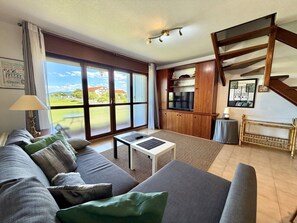 Un salon avec un canapé, une table basse et un téléviseur. On y trouve un escalier et une vue sur l’extérieur.