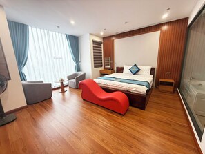 A modern hotel room with a wooden floor, a bed with a blue and white bedspread, a red armchair, a grey armchair, and a small table with a fan.