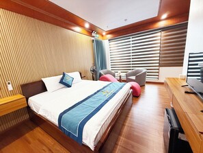 A hotel room with a wooden headboard, a bed with a blue and white bedspread, a desk with a television, and a window with blinds.