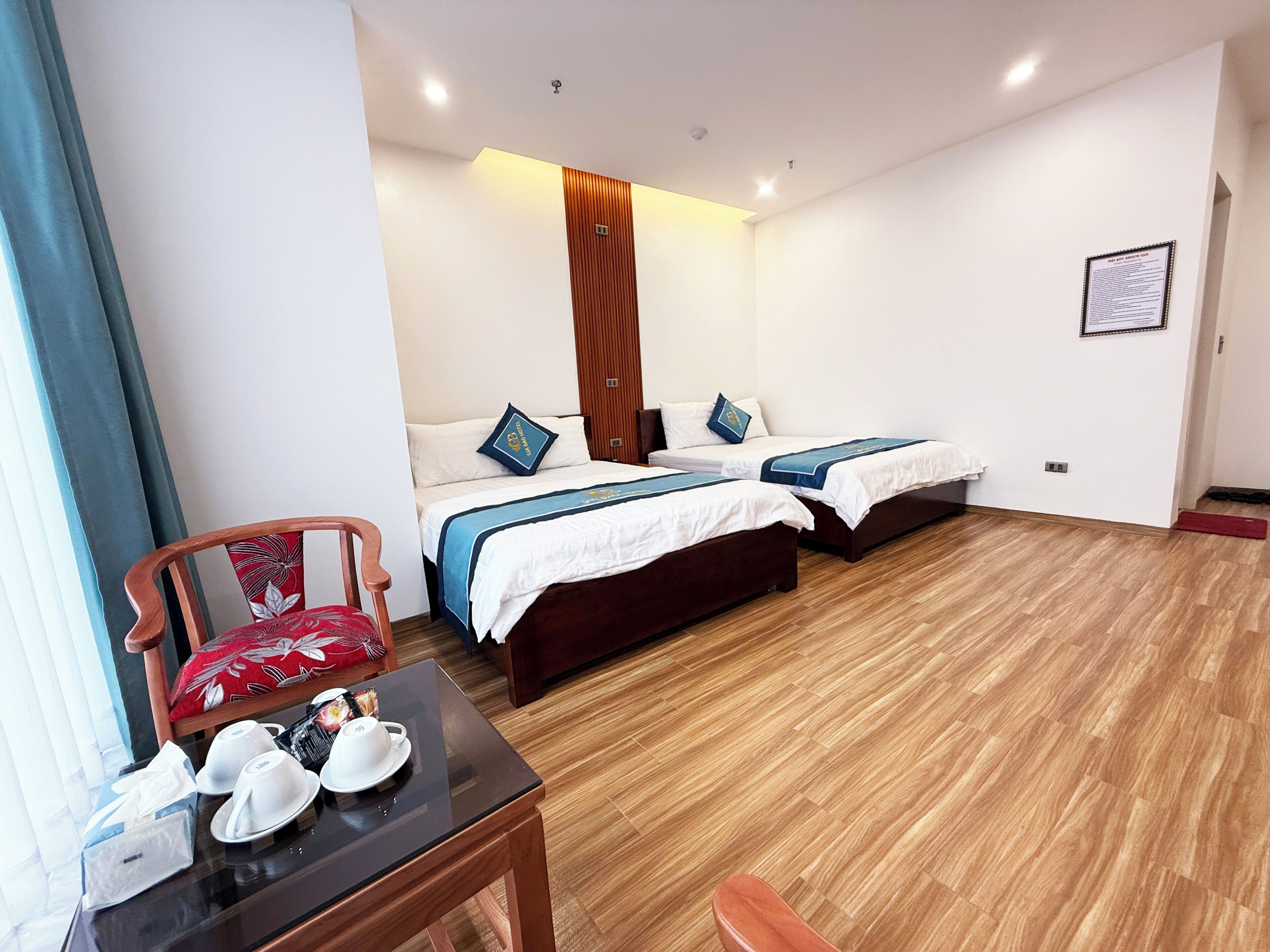 A hotel room with two beds, a wooden floor, a small table with a chair, and a framed certificate on the wall.