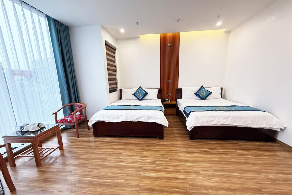 A hotel room with two beds, wooden flooring, a small table with a chair, and a window with curtains.