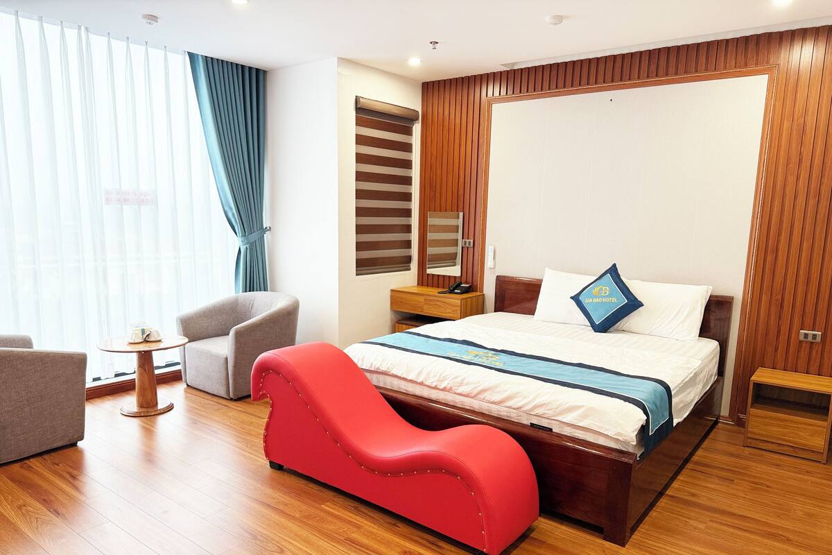 A hotel room with a bed, a red sofa, a wooden floor, a window with curtains, and a wooden panel wall.
