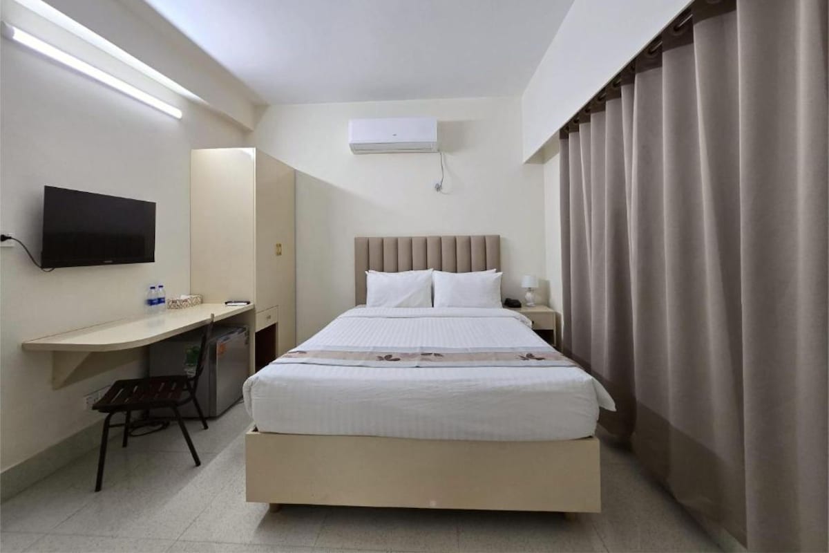 A hotel room with a bed, a desk with a chair, a television, an air conditioning unit, and curtains.