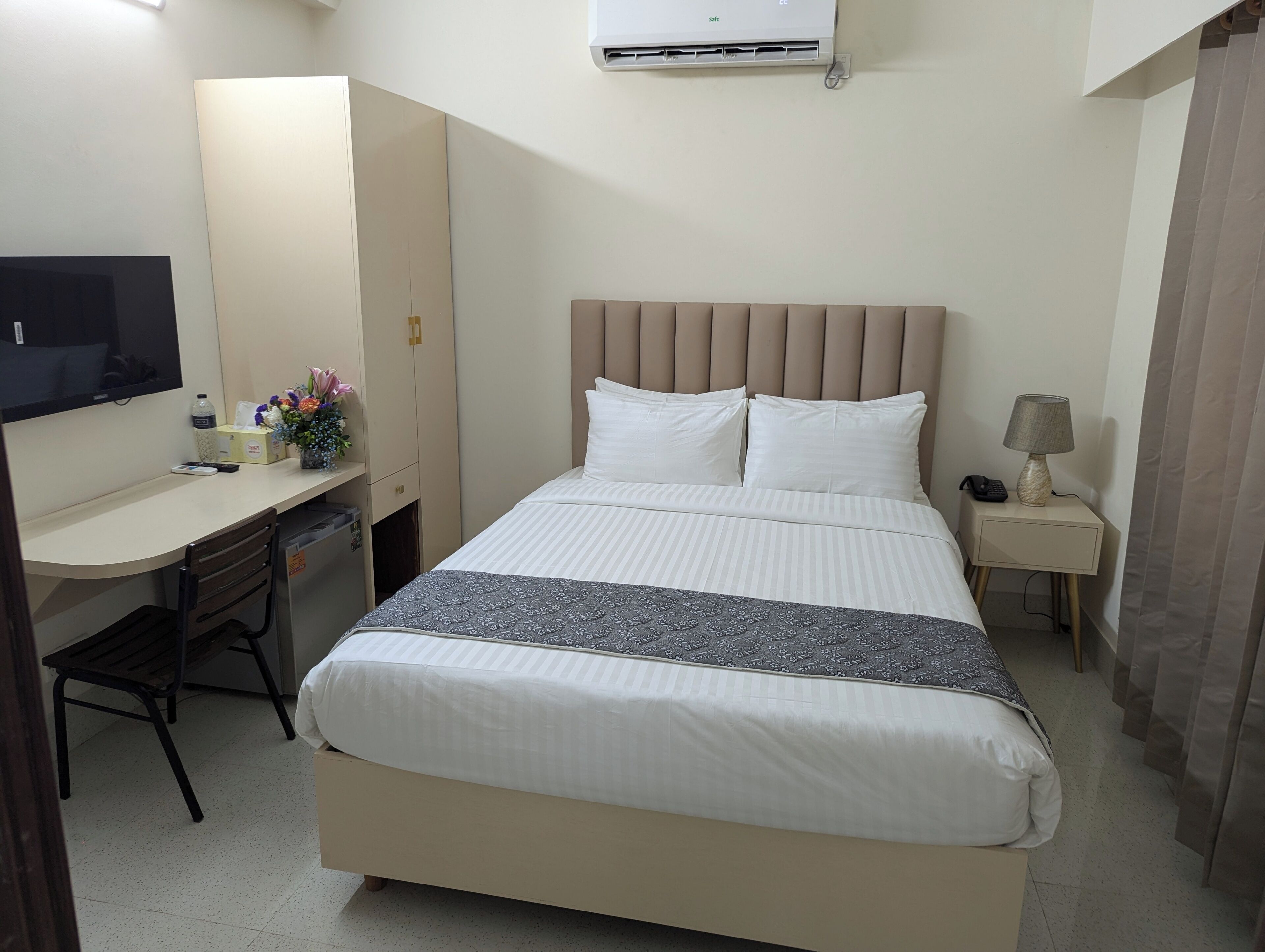 A hotel room with a bed, a desk, a chair, a TV, a bedside table with a lamp, and an air conditioning unit.