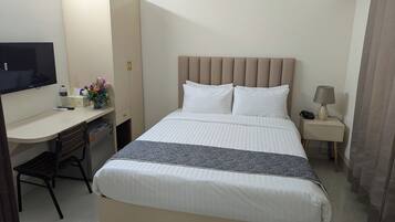 A hotel room with a bed, a desk, a chair, a TV, a bedside table with a lamp, and an air conditioning unit.