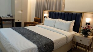 A hotel room with a large bed, a desk with a chair, a wardrobe, and a nightstand with a lamp and flowers. - Royal Regent Suites Dhaka (Dhaka)