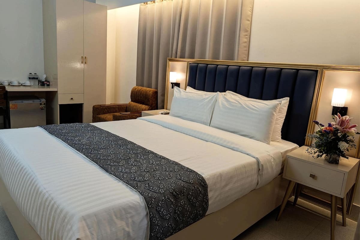 A hotel room with a large bed, a desk with a chair, a wardrobe, and a nightstand with a lamp and flowers.
