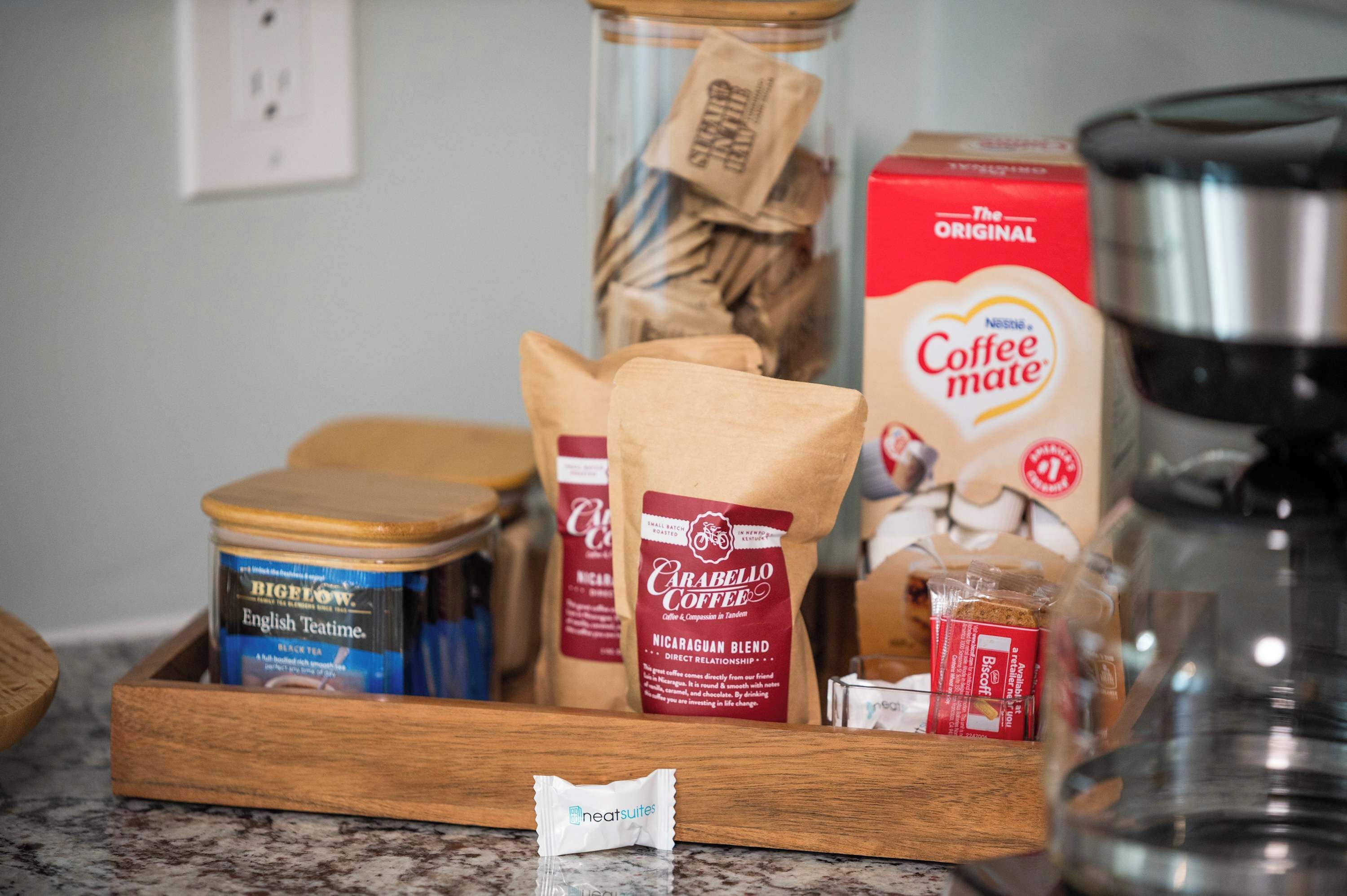 Un comptoir de cuisine où l’on trouve divers produits à base de café, notamment une boîte de café « The Original », un bocal de café en grains « Carabello Coffee », un contenant de thé anglais « Bigelow » et une boîte de sachets de thé « Heartsuites ».