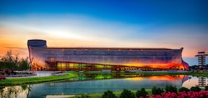 A large, modern building with a unique curved roof, situated next to a calm body of water, reflecting the structure.