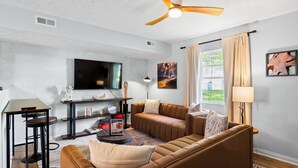 A modern living room with a sofa, coffee table, and TV. There is a ceiling fan, a window with curtains, and a lamp. - 3BR Condo Near Ark Encounter, 8 Guests, Parking (Williamstown)