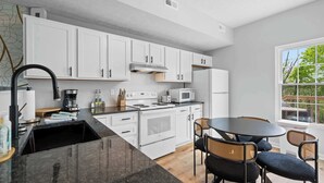 Una cocina moderna con gabinetes blancos, encimera negra y una zona de comedor con sillas de mimbre.