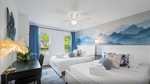 Habitación de hotel con dos camas, un escritorio, un ventilador de techo y un mural de montañas.
