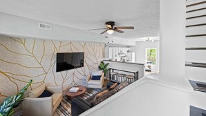 Una sala de estar moderna con sofá, sillón y mesa de centro, un televisor de pantalla plana montado en la pared y un ventilador de techo.