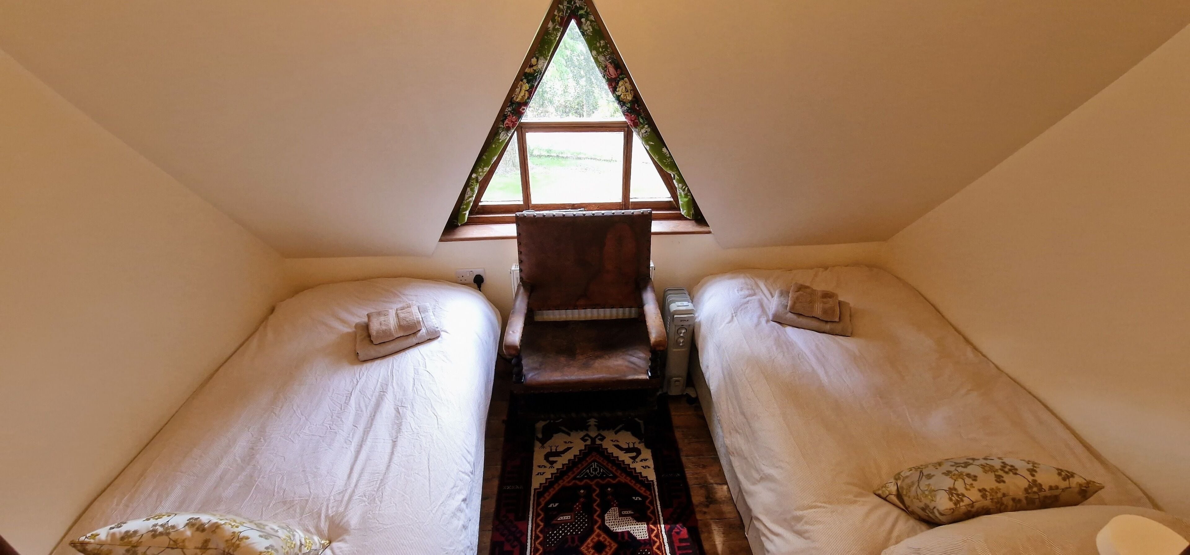 A small, cozy room with two single beds, a chair, and a window with a floral curtain.