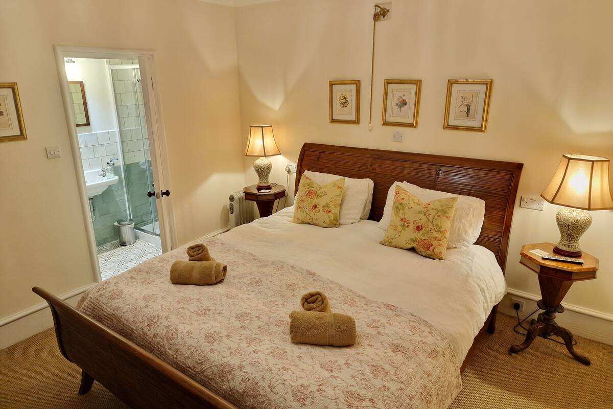 A hotel room with a large bed, two bedside tables with lamps, a wooden headboard, floral-patterned bedding, and a small bathroom visible through an open door.