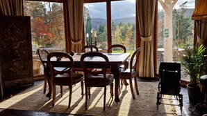 Un espace repas comprenant une table et des chaises, de grandes fenêtres avec des rideaux, et une vue sur les arbres et les montagnes.