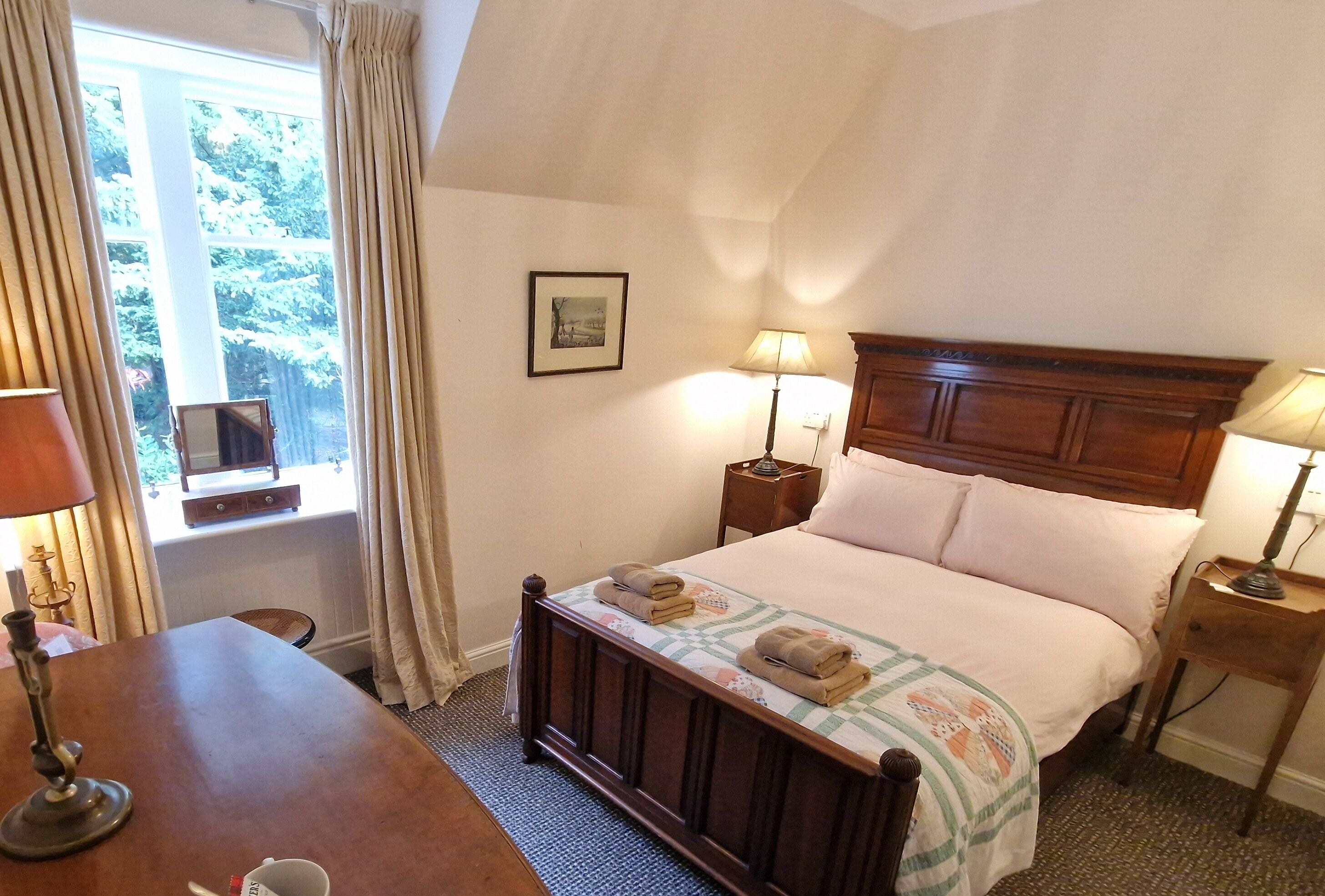 A bedroom with a wooden bed, two bedside lamps, a window with curtains, a framed picture on the wall, and a small table with a lamp.