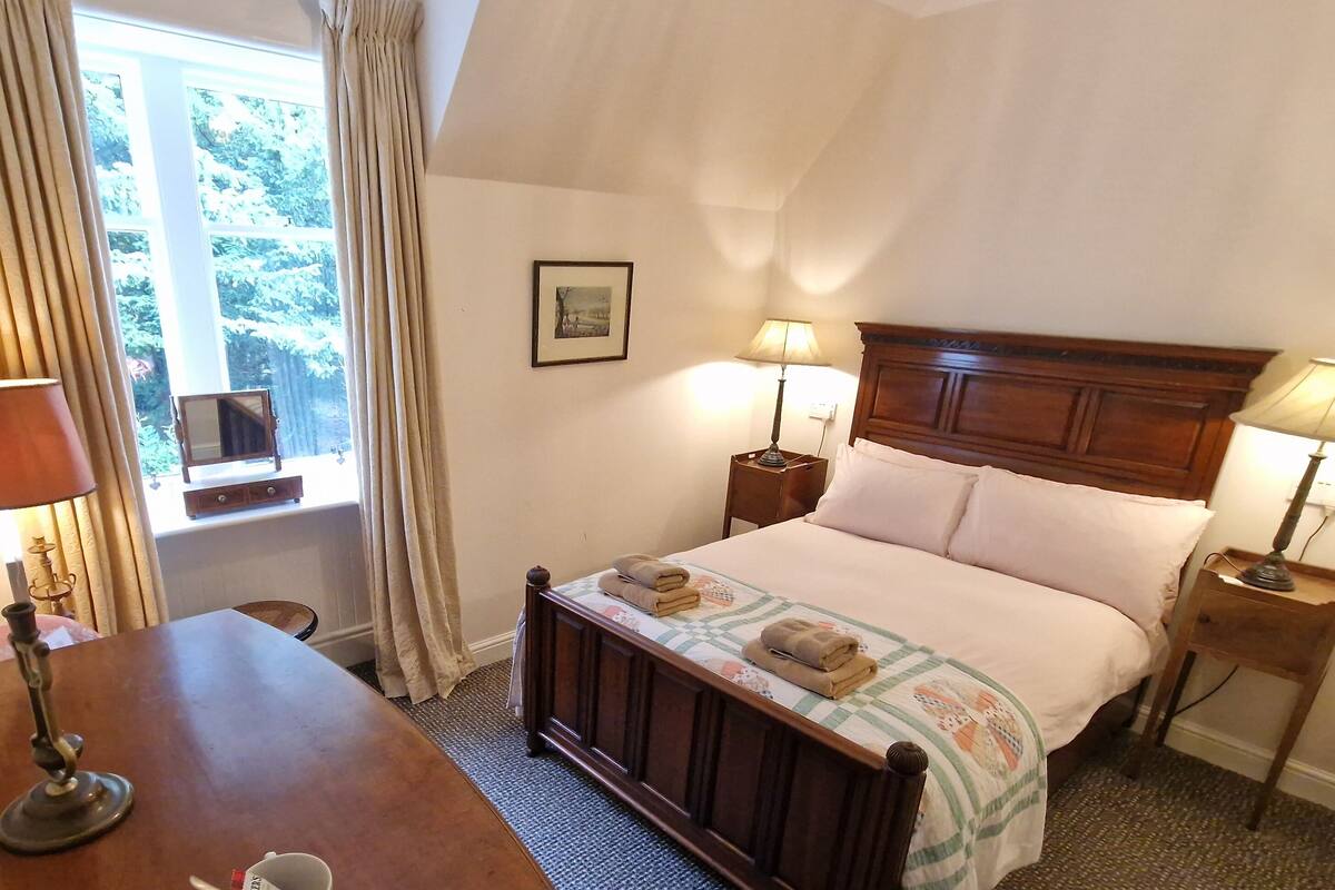 A bedroom with a wooden bed, two bedside lamps, a window with curtains, a framed picture on the wall, and a small table with a lamp.