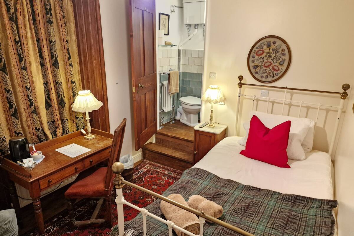 A hotel room with a bed, a desk with a lamp, a chair, a small table, a red pillow, a patterned rug, and a bathroom visible through an open door.
