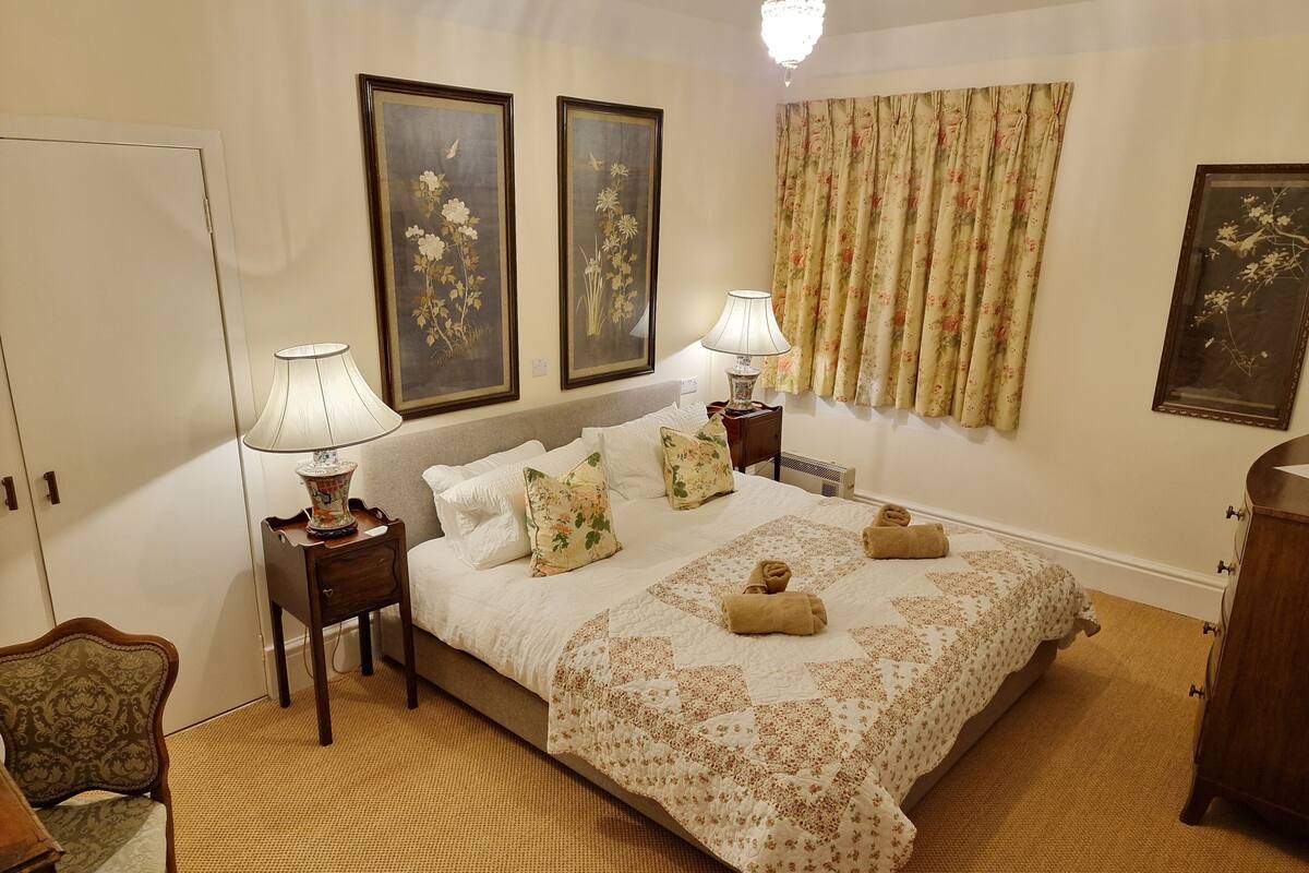A neatly arranged bedroom with a large bed, two bedside lamps, floral curtains, and framed artwork on the walls.