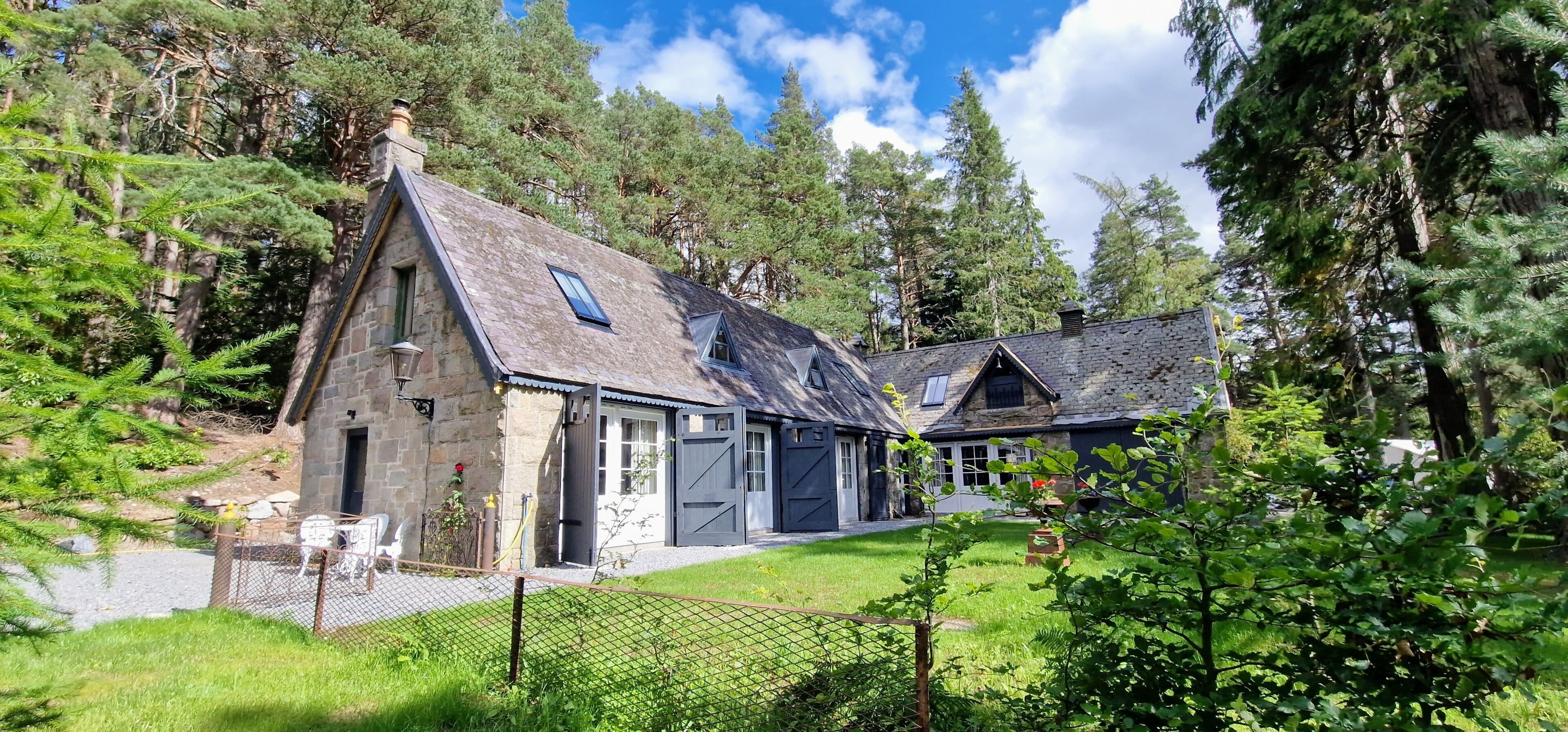 A stone cottage with a sloped roof, a fenced yard, and a view of a forested area.