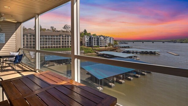 A balcony with a view of a sunset over water, a building in the background, and a table with chairs. - Immaculate Lands End #735 3Bed/2Bath New Remodel! (Osage Beach)