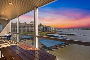 A balcony with a view of a sunset over water, a building in the background, and a table with chairs.