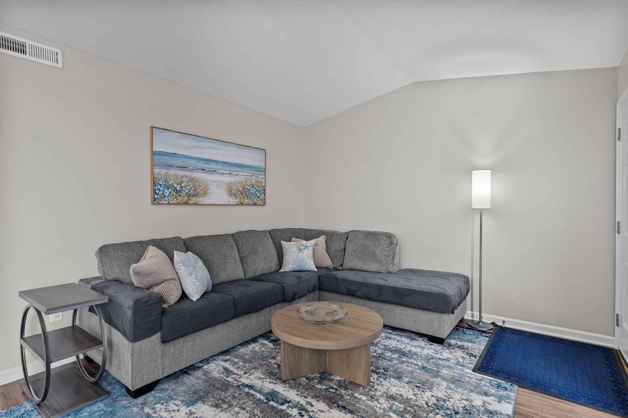 Een moderne woonkamer met een grijze modulaire bank, een klein bijzettafeltje, een ronde salontafel, een blauw tapijt met patroon en een schilderij van een strandgezicht.