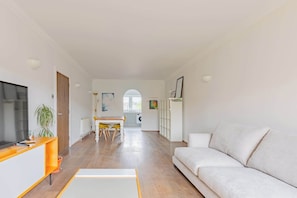 Uma sala de estar moderna com sofá, televisão, área de jantar com cadeiras e cozinha ao fundo.