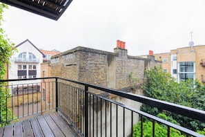 Uma varanda com piso de madeira e guarda-corpo preto, com vista para um prédio de tijolos com chaminés vermelhas e uma casa branca com varanda ao fundo.