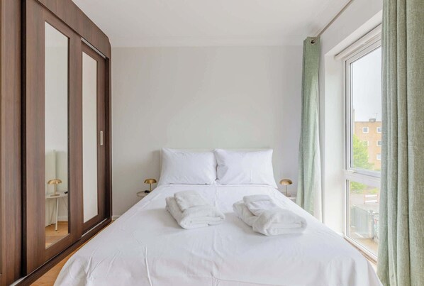 A neatly made bed with white linens and two folded towels, a large mirror, and a window with curtains.