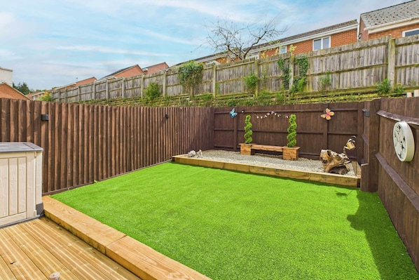 A backyard with artificial grass, a wooden deck, a wooden fence, and a bench.