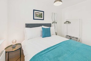 A neatly made bed with white and blue bedding, a small bedside table with a lamp, a framed picture on the wall, and a radiator.