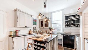 A modern kitchen with white cabinetry, a central island with wine storage, pendant lighting, and a view of the city through large windows. - Fabulous Solihull Luxury Designer Apartment 3BR (West Midlands)