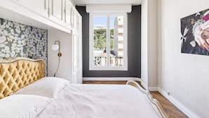 A bedroom with a tufted headboard, a large window with a view of buildings, and a painting on the wall.