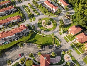 An aerial view of a residential area with a central roundabout, houses with red roofs, and a circular park with palm trees.