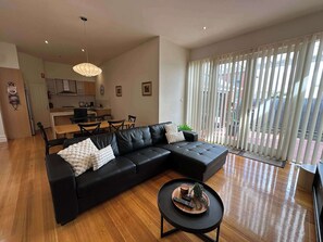 A modern living room with a sectional sofa, a dining table, and chairs. There is a kitchen in the background with cabinets and a window. - Central Geelong Retreat, Sleeps 6! (Geelong)