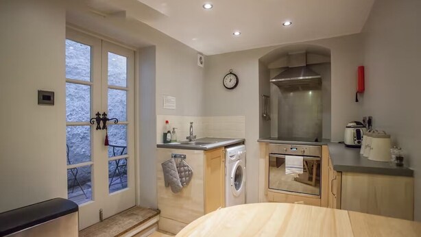 A compact kitchen with a washing machine, oven, and a clock on the wall. - Tide & Teacups (Brighton)