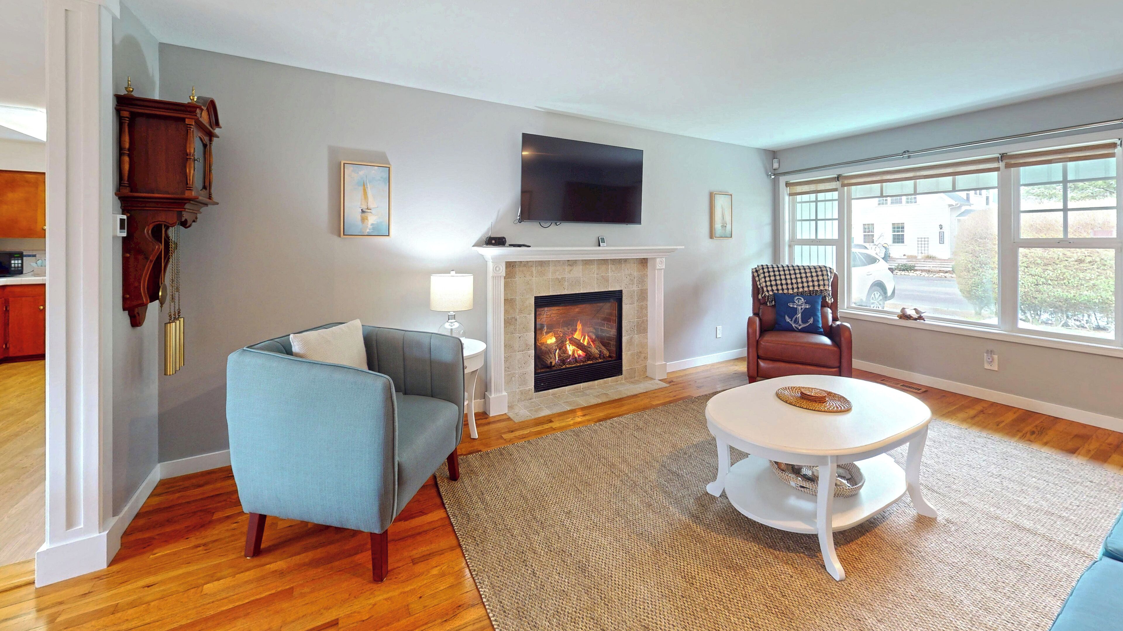 A living room with a fireplace, a television mounted above it, a blue armchair, a round white coffee table, and a view through large windows.