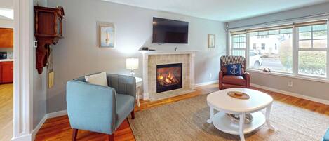 A living room with a fireplace, a television mounted above it, a blue armchair, a round white coffee table, and a view through large windows.