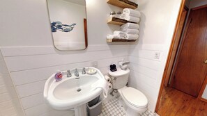 A bathroom with a white sink, toilet, and a mirror. There are towels on a shelf above the toilet. - Tobey's Treehouse Oasis at Seaside Beach (Seaside)