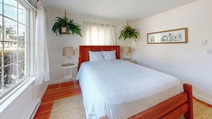 A bedroom with a large bed, two bedside tables with lamps, a window with curtains, and two hanging plants. - Edgewood Escape at Seaside Beach (Seaside)