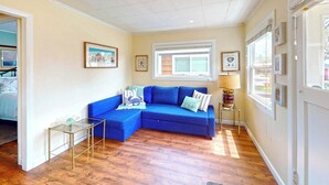 A living room with a blue sectional sofa, a small side table, and a lamp. There are framed pictures on the walls and a window with blinds. - Sandpiper Cottage at Seaside Beach (Seaside)