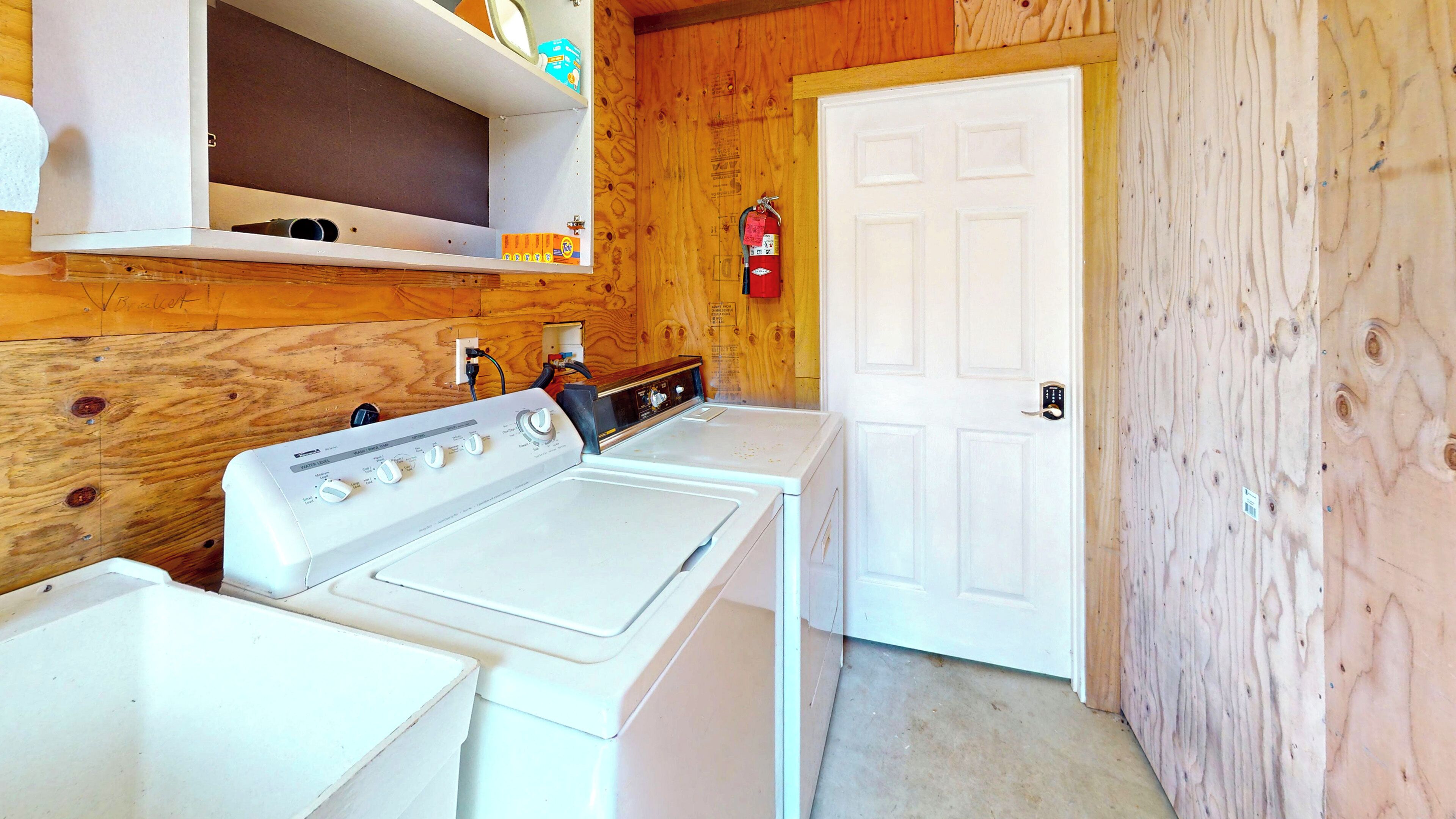 A laundry room with a white washer and dryer, wooden walls, a white door, and a fire extinguisher mounted on the wall.
