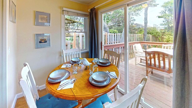 A dining area with a wooden table set for four, white chairs, and a view of the deck and trees outside. - Sandpiper Cottage at Seaside Beach (Seaside)
