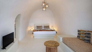 A modern, minimalist bedroom with a large bed, a TV, a small wooden stool, and a sofa. - Amphora Meltemi Seaview Gem w/ Private Jacuzzi (Akrotiri)